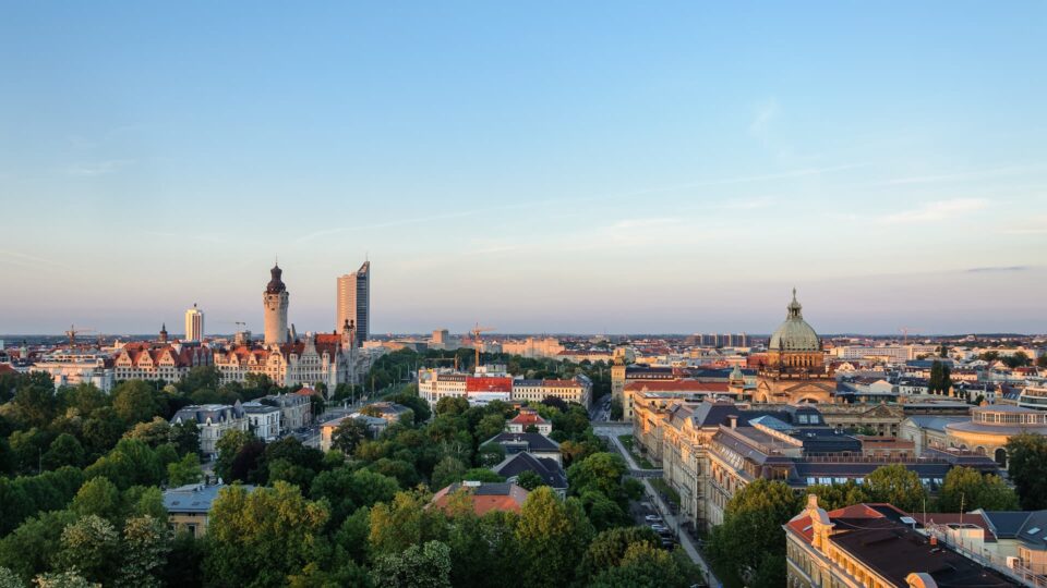 Ein Panoramablick auf Leipzig bei Sonnenuntergang zeigt eine Mischung aus modernen und historischen Gebäuden, darunter ein hohes Hochhaus und eine historische Kuppelkonstruktion. Im Vordergrund sind grüne Bäume und Wohnhäuser zu sehen, während der Himmel teilweise klar ist und sanftes Abendlicht bietet.