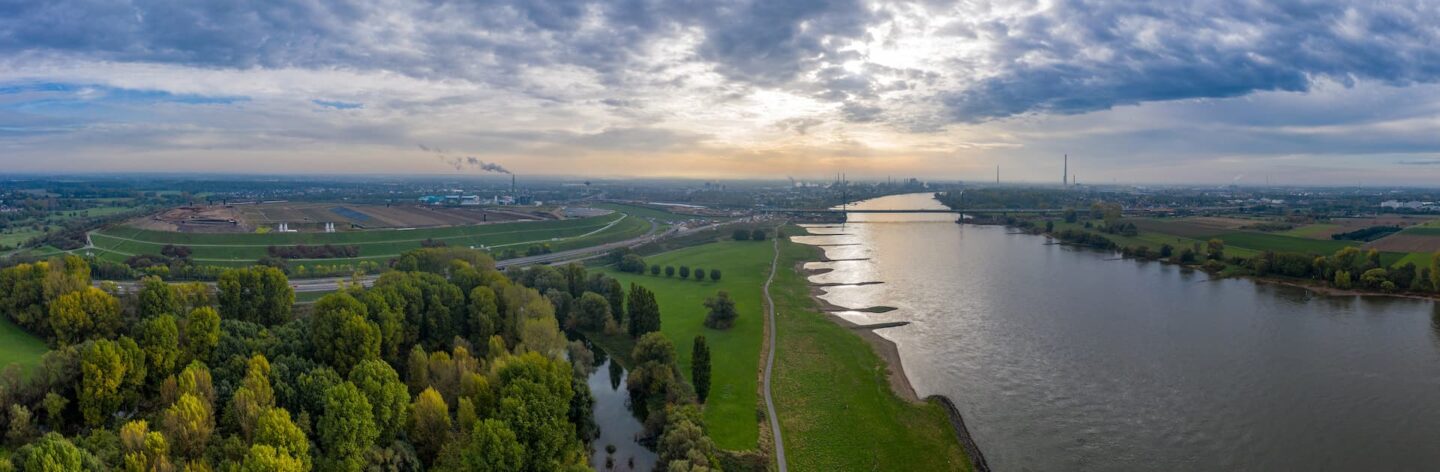 Eine Panorama-Luftaufnahme eines Flusses, der auf beiden Seiten von üppigen grünen Bäumen und Feldern gesäumt ist. Der Himmel darüber ist mit einer Mischung aus Wolken bedeckt und am fernen Horizont ist die Stadt- und Industrielandschaft von Leverkusen zu sehen, wobei einige Schornsteine sichtbar sind.