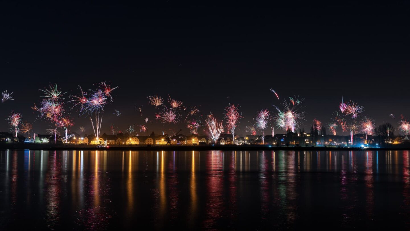 Ein Nachthimmel voller bunter Feuerwerke über der Stadt Leverkusen. Darunter erzeugen die Lichter der Gebäude entlang der Uferpromenade Spiegelungen im Wasser. Die Szene fängt die hellen Explosionen und die friedliche, beleuchtete Spiegelung auf der ruhigen Wasseroberfläche ein.