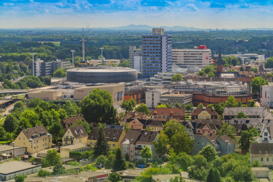 Luftaufnahme der Stadtlandschaft von Leverkusen, die im Vordergrund eine Mischung aus Wohnhäusern und im Hintergrund moderne Geschäftsgebäude, darunter ein kreisförmiges Gebäude, zeigt. Am Horizont sind Grünflächen und entfernte Hügel unter einem blauen, teilweise bewölkten Himmel zu sehen.