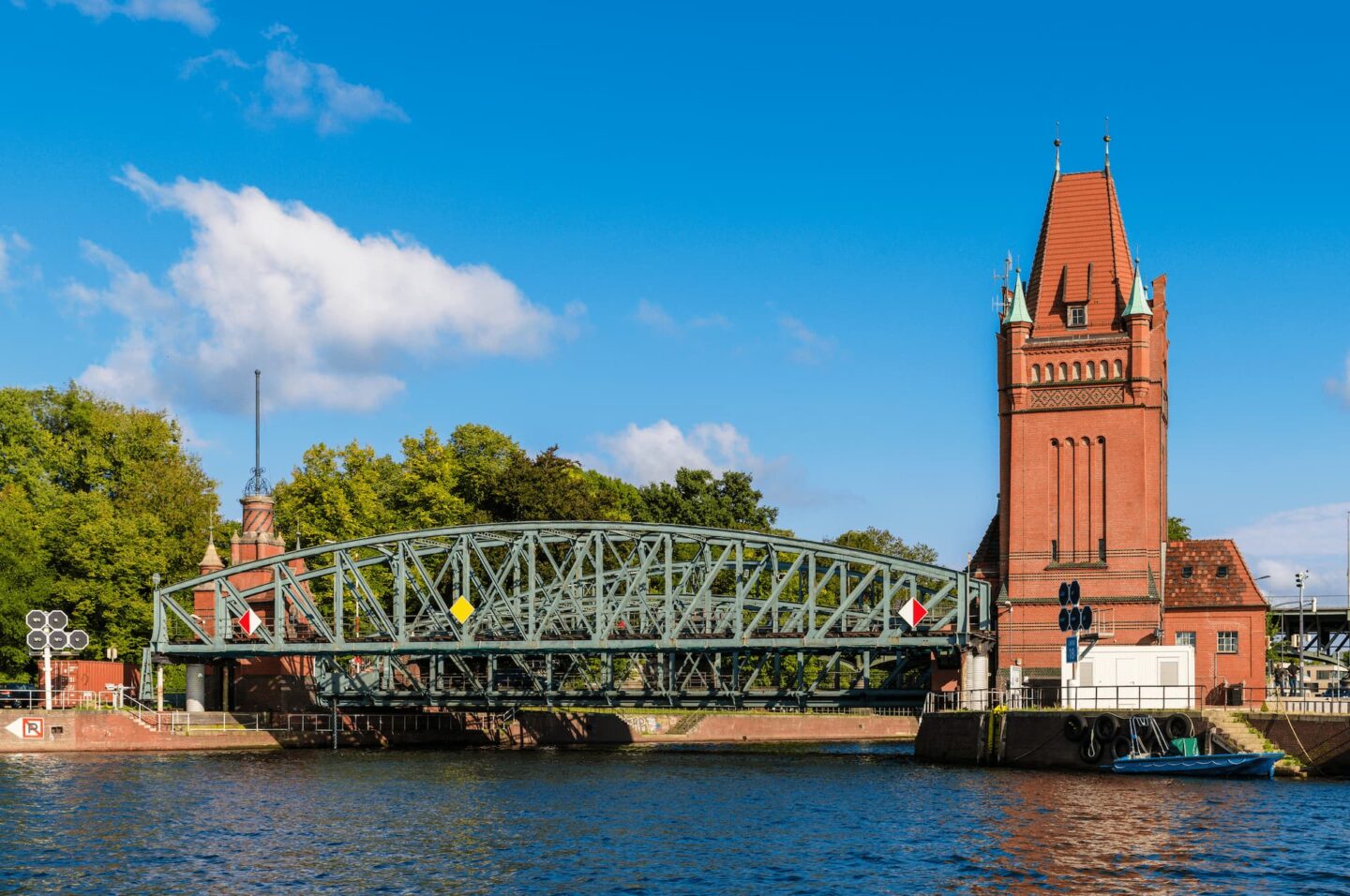 Eine grüne Zugbrücke aus Stahl überspannt einen Fluss neben einem hohen Gebäude aus rotem Backstein mit steilem Spitzdach. Die malerische Aussicht liegt in Lübeck, Deutschland, vor einer Kulisse aus Bäumen und einem strahlend blauen Himmel mit vereinzelten Wolken – perfekt für Tourismusbegeisterte.