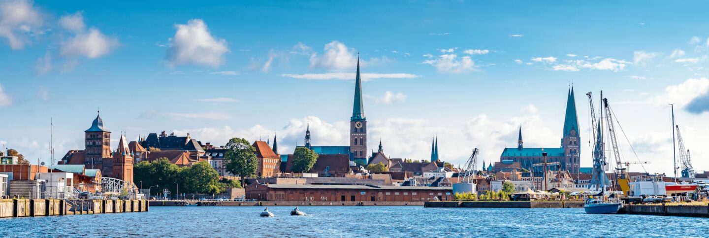 Panoramablick auf die Küstenstadt Lübeck, Deutschland, mit einer Ansammlung historischer Gebäude und Kirchtürme unter einem blauen Himmel mit vereinzelten Wolken. Im Vordergrund liegen Boote und Yachten vor Anker, und das Wasser ist ruhig und spiegelt die malerische Skyline wider – ein perfekter Ort für den Tourismus.