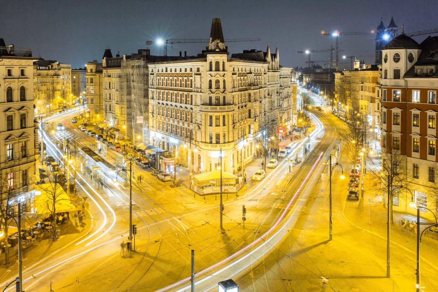 Eine nächtliche Stadtansicht in Magdeburg, Deutschland, mit beleuchteten Straßen und regem Verkehr, eingefangen durch Lichtspuren. Die Szene zeigt historische Gebäude, darunter ein großes zentrales Gebäude mit Spitzdach, umgeben von Straßenlaternen, Fußgängern und sichtbaren Baukränen im Hintergrund.