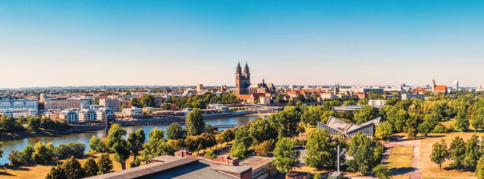 Ein Panoramablick auf die Stadtlandschaft Magdeburgs mit einem Fluss, üppigen grünen Bäumen und historischen Gebäuden. Die Skyline wird vom großen Dom mit seinen zwei Türmen dominiert, während im Hintergrund moderne Gebäude unter einem klaren, blauen Himmel stehen. Perfekt für jeden Reiseführer oder Tourismusliebhaber.