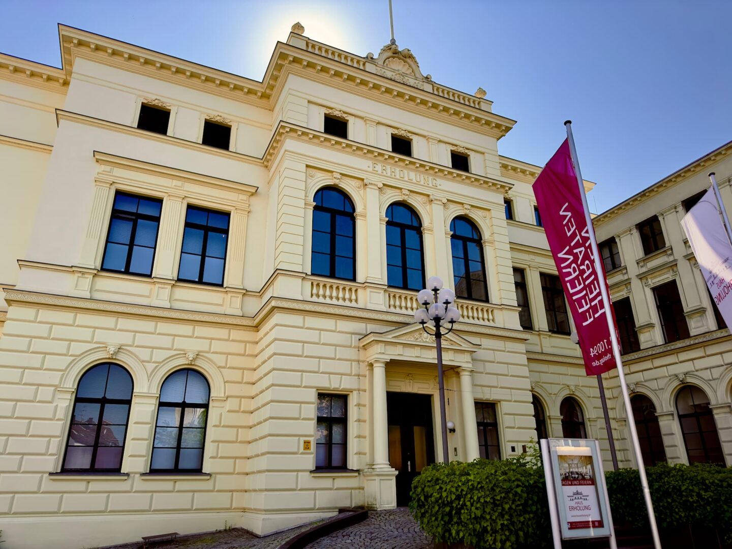 Ein historisches cremefarbenes Gebäude mit Bogenfenstern und kunstvollen architektonischen Details steht stolz in Mönchengladbach. Vorne hängen Banner und ein Laternenpfahl schmückt den Eingang. Ein heller, klarer Himmel dient als Kulisse, während ein kleiner Garten mit viel Grün der Vorderseite Charme verleiht.