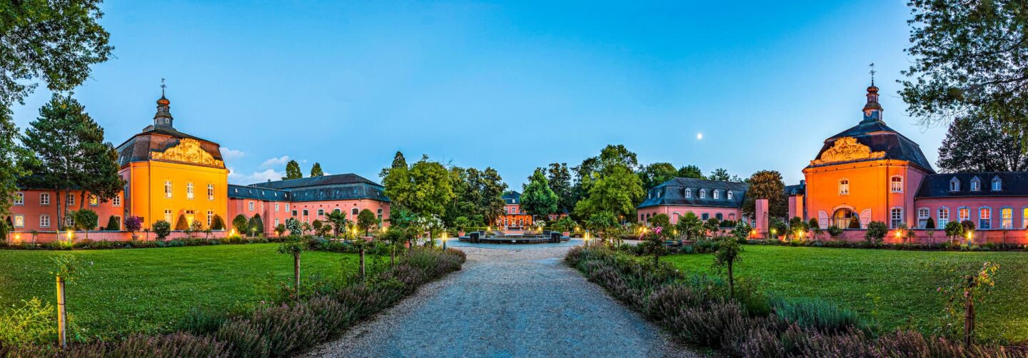 Panoramablick auf einen malerischen Schlosskomplex in Mönchengladbach am frühen Abend. Das Bild zeigt zwei orangefarbene Gebäude mit Kuppeldächern auf beiden Seiten eines Weges, der zu einem zentralen Brunnen führt, der von Grün und Bäumen unter einem klaren blauen Himmel mit sichtbarem Mond umgeben ist.