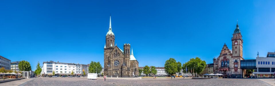 Ein Panoramablick auf den Stadtplatz von Mönchengladbach zeigt eine große historische Kirche mit einem hohen Turm und einem Uhrturm in der Mitte. Flankiert von anderen architektonischen Gebäuden und gesäumt von Bäumen bietet der Platz einen offenen, gepflasterten Raum unter einem klaren blauen Himmel.