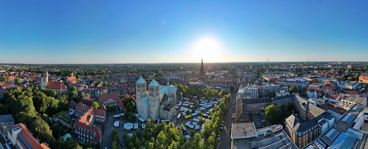 Ein Panorama-Luftbild von Münster bei Sonnenuntergang offenbart eine atemberaubende Stadtlandschaft mit einer Mischung aus historischen und modernen Gebäuden. Nahe dem Zentrum steht ein markanter Dom, umgeben von dichter städtischer Bebauung, Grünflächen und einem geschäftigen Marktgebiet. Der Himmel ist klar und die Sonne steht tief am Horizont.