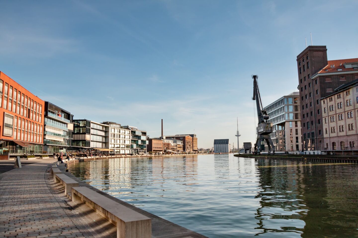 Blick auf eine Uferszene in Münster mit einem Kanal, der links von modernen Gebäuden und rechts von Gebäuden im Industriestil in einem Hafengebiet gesäumt ist. Unter einem klaren blauen Himmel sind ein großer Kran und ein entfernter Fernsehturm zu sehen. Der Kopfsteinpflasterweg säumt das Wasser.