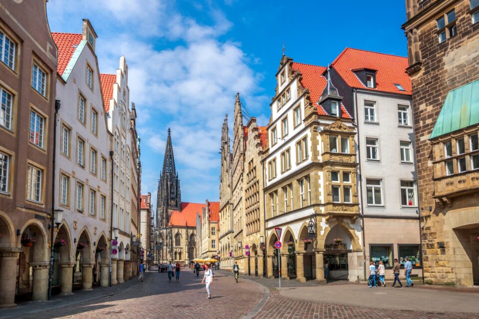 Eine belebte Straße in einer europäischen Stadt mit historischer Architektur, mit Gebäuden mit roten Ziegeldächern, einem hohen Kirchturm in der Ferne und Menschen, die die Kopfsteinpflasterstraße entlanggehen. Der Himmel ist blau mit vereinzelten Wolken und erinnert an malerische Münstertage.