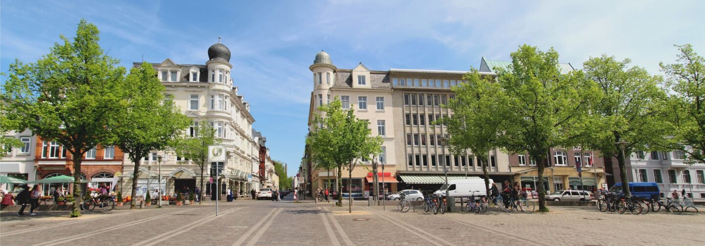 Eine breite Straße, gesäumt von grünen Bäumen, Straßencafés und verschiedenen Gebäuden in unterschiedlichen Architekturstilen, prägt die charmante Promenade von Neumünster. Menschen sitzen in den Cafés, während ein paar Fahrräder und Autos entlang der Straße unter einem klaren blauen Himmel parken.