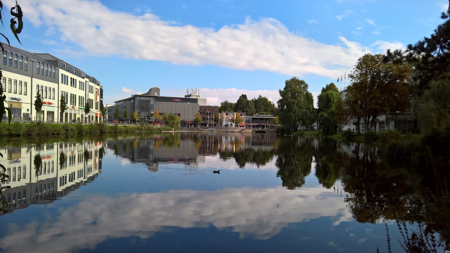 Ein ruhiger See mit Spiegelungen der umliegenden Gebäude und Grünflächen unter einem strahlend blauen Himmel mit vereinzelten Wolken in Neumünster. Eine einsame Ente schwimmt in der Nähe der Mitte und trägt zur friedlichen Atmosphäre bei. Die modernen Gebäude und üppigen Bäume sorgen für ein Gleichgewicht zwischen städtischen und natürlichen Elementen.