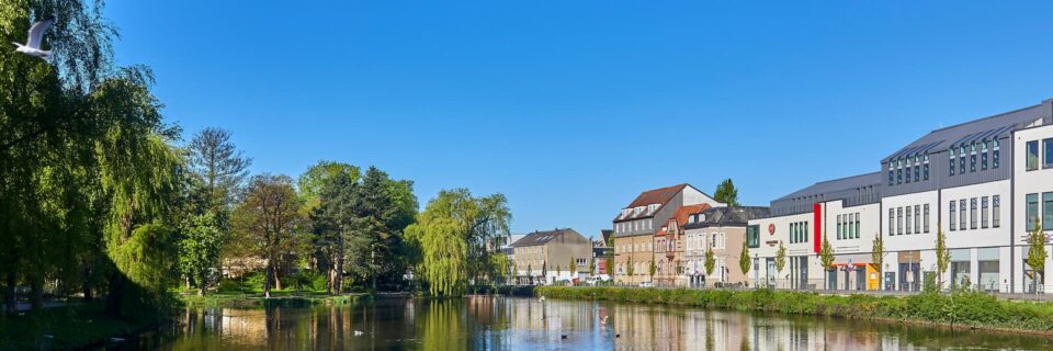 Ein malerischer Blick auf einen ruhigen Fluss in Neumünster, flankiert von Bäumen auf der linken und Gebäuden auf der rechten Seite. Der klare blaue Himmel spiegelt sich im Wasser und man sieht ein paar Vögel fliegen. Üppiges Grün und farbenfrohe Fassaden der Gebäude tragen zur ruhigen Atmosphäre bei.