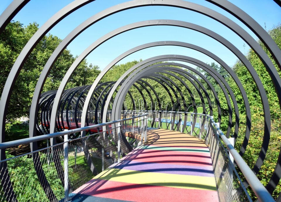 Eine Fußgängerbrücke in Oberhausen besteht aus einer Reihe von Metallbögen, die gemusterte Schatten auf einen regenbogenfarbenen Gehweg werfen. Der Weg ist von üppigen grünen Bäumen unter einem klaren blauen Himmel umgeben.