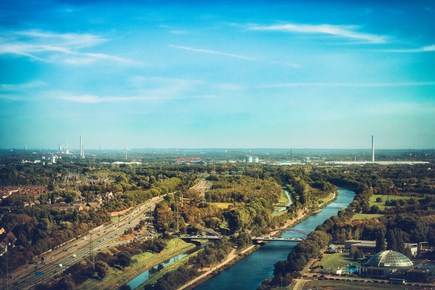 Luftaufnahme einer Landschaft mit einem Fluss, der sich durch Waldgebiete und Parks in Oberhausen schlängelt. Die Szene umfasst Brücken über den Fluss, Straßen und einige Industriegebäude am Horizont unter einem klaren blauen Himmel.