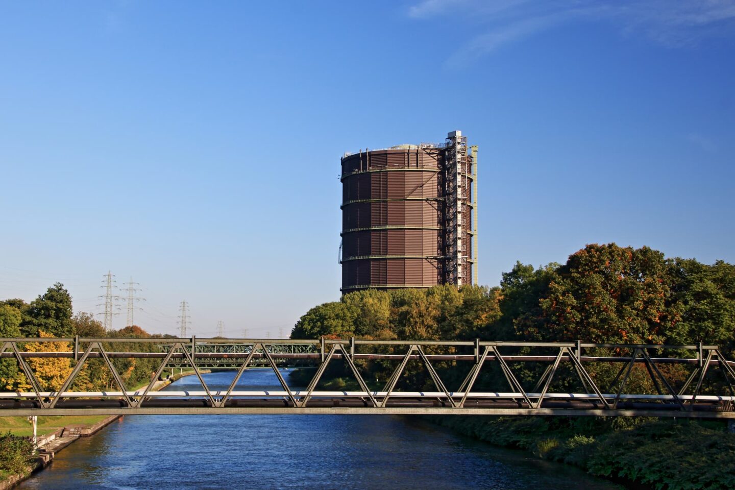 Ein hohes, zylindrisches Industriegebäude in Oberhausen, Deutschland, steht vor einem klaren blauen Himmel, umgeben von grünen Bäumen. Im Vordergrund überspannt eine Metallbrücke einen Wasserweg, an dessen Ufern weitere Bäume stehen. In der Ferne sind schwach Stromleitungen und Türme zu erkennen.