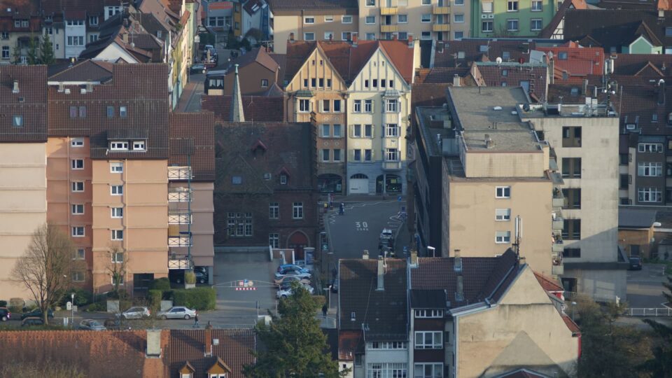 Luftaufnahme von Pforzheim, einer Stadt in Deutschland, die für ihre Schmuckindustrie bekannt ist. Sie zeigt ein europäisches Stadtviertel mit dicht gedrängten Gebäuden in einer Vielzahl von Architekturstilen und Farben. Straßen kreuzen sich zwischen den Gebäuden, und entlang der Straßen und auf kleinen Parkplätzen sind mehrere geparkte Autos zu sehen.