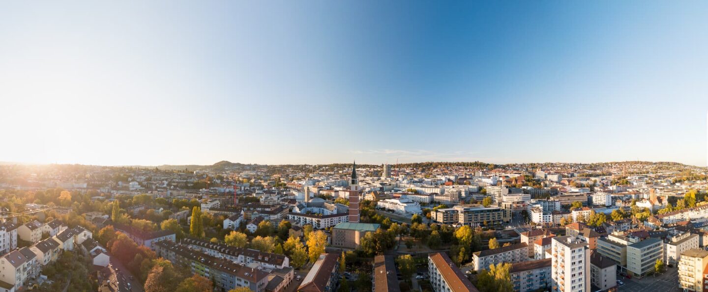 Ein Panoramablick auf Pforzheim unter einem klaren blauen Himmel. Die Stadtlandschaft zeichnet sich durch zahlreiche Wohn- und Geschäftsgebäude aus und ist von Grünflächen und Bäumen umgeben. Der Horizont ist sichtbar, in der Ferne sind Hügel zu sehen und die Sonne wirft ein helles Licht auf die Szene.