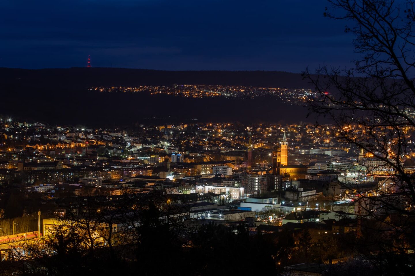 Eine nächtliche Stadtlandschaft mit beleuchteten Gebäuden und Straßen. Die Lichter bilden einen lebendigen Kontrast zum dunklen Himmel. In der Ferne ist ein hohes Gebäude mit einem deutlich leuchtenden Licht zu sehen, zusammen mit einem Turm auf einem entfernten Hügel, der ein rotes Licht ausstrahlt und an die Skyline von Pforzheim erinnert.