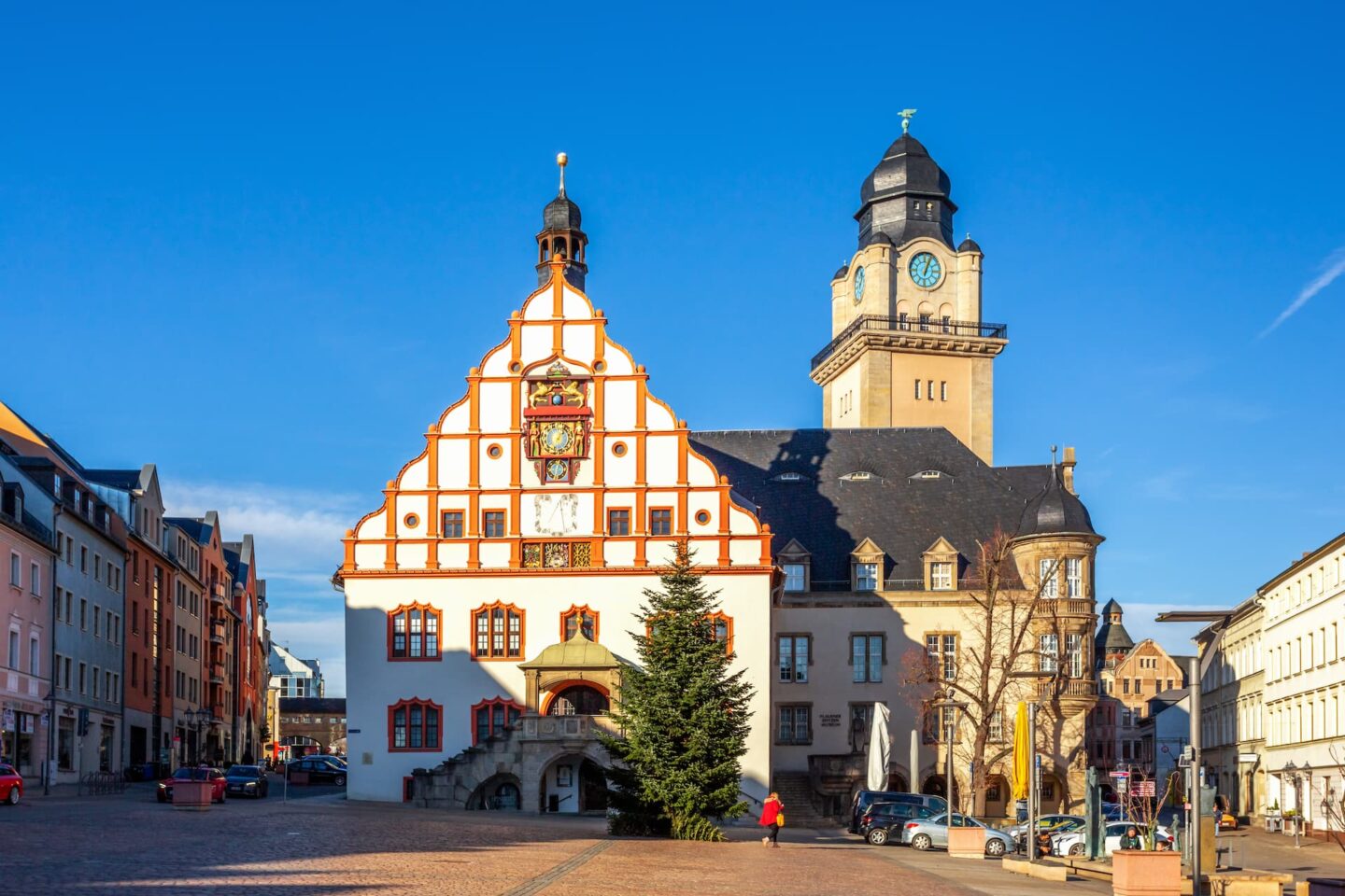 Ein malerischer Stadtplatz in Plauen mit historischen Gebäuden. Das zentrale Gebäude verfügt über einen hohen Uhrturm, steile Giebeldächer und verzierte Fenster. Vorne steht ein großer, geschmückter Weihnachtsbaum und eine Person in einem hellen Mantel geht daneben.