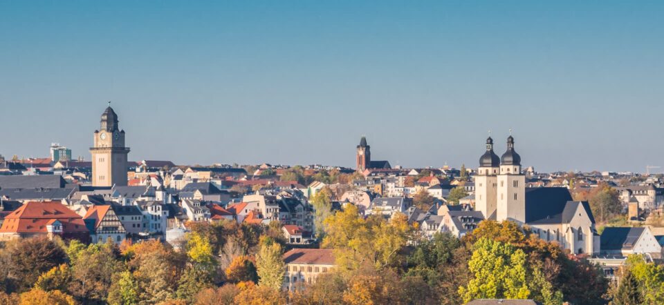 Panoramablick auf die historische europäische Stadt Plauen mit markanten Uhrtürmen und einer Kirche mit zwei Türmen. Die Stadt ist von buntem Herbstlaub unter einem klaren blauen Himmel umgeben. Die Gebäude weisen eine Mischung aus klassischem und modernem Architekturstil auf.