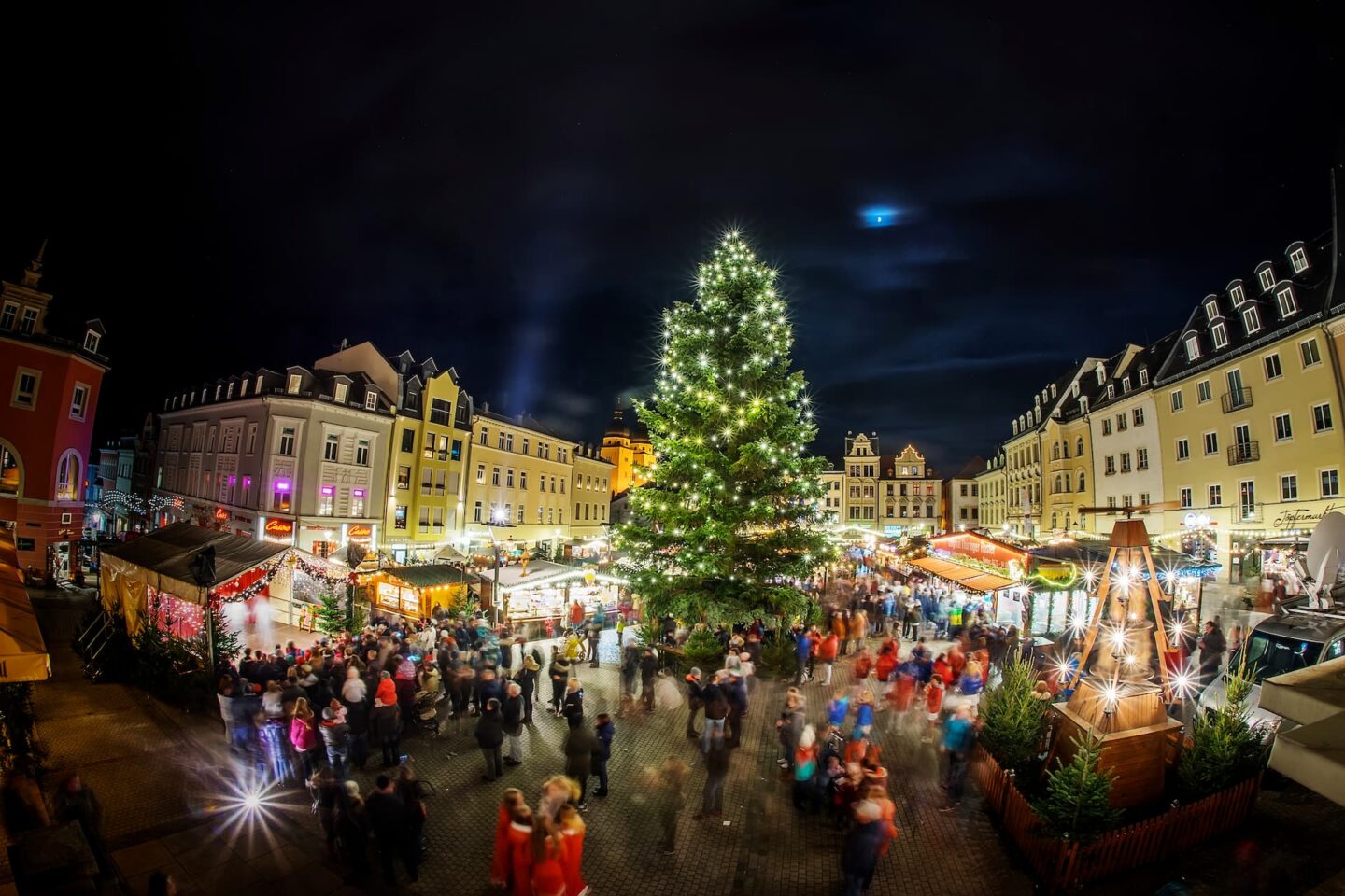 Ein festlicher Weihnachtsmarkt bei Nacht, beleuchtet von bunten Lichtern. Im Mittelpunkt steht ein großer, geschmückter Weihnachtsbaum, umgeben von geschäftigen Menschenmengen und zahlreichen Ständen. Historische Gebäude rahmen das Bild in Plauen ein und tragen zur bezaubernden Weihnachtsstimmung bei.