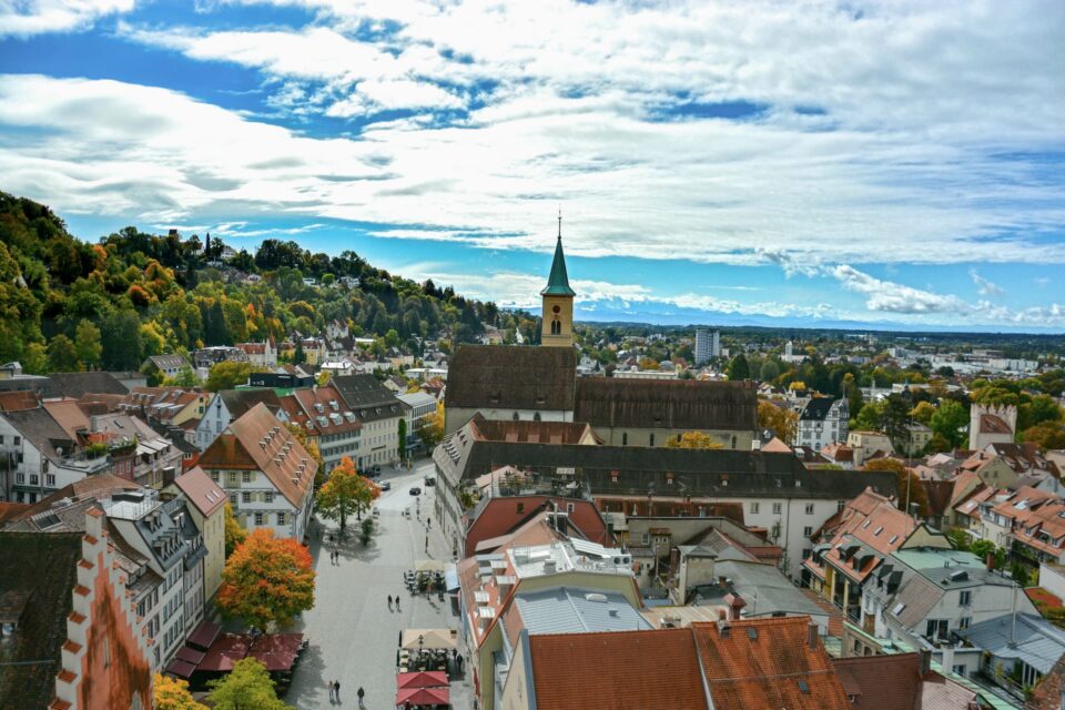 Eine Luftaufnahme der europäischen Stadt Ravensburg zeigt eine zentrale Kirche mit einem hohen Turm, umgeben von Gebäuden mit roten Dächern. Die Szene zeigt Menschen, die auf den Straßen spazieren gehen, und im Hintergrund üppige grüne Hügel unter einem teilweise bewölkten Himmel.