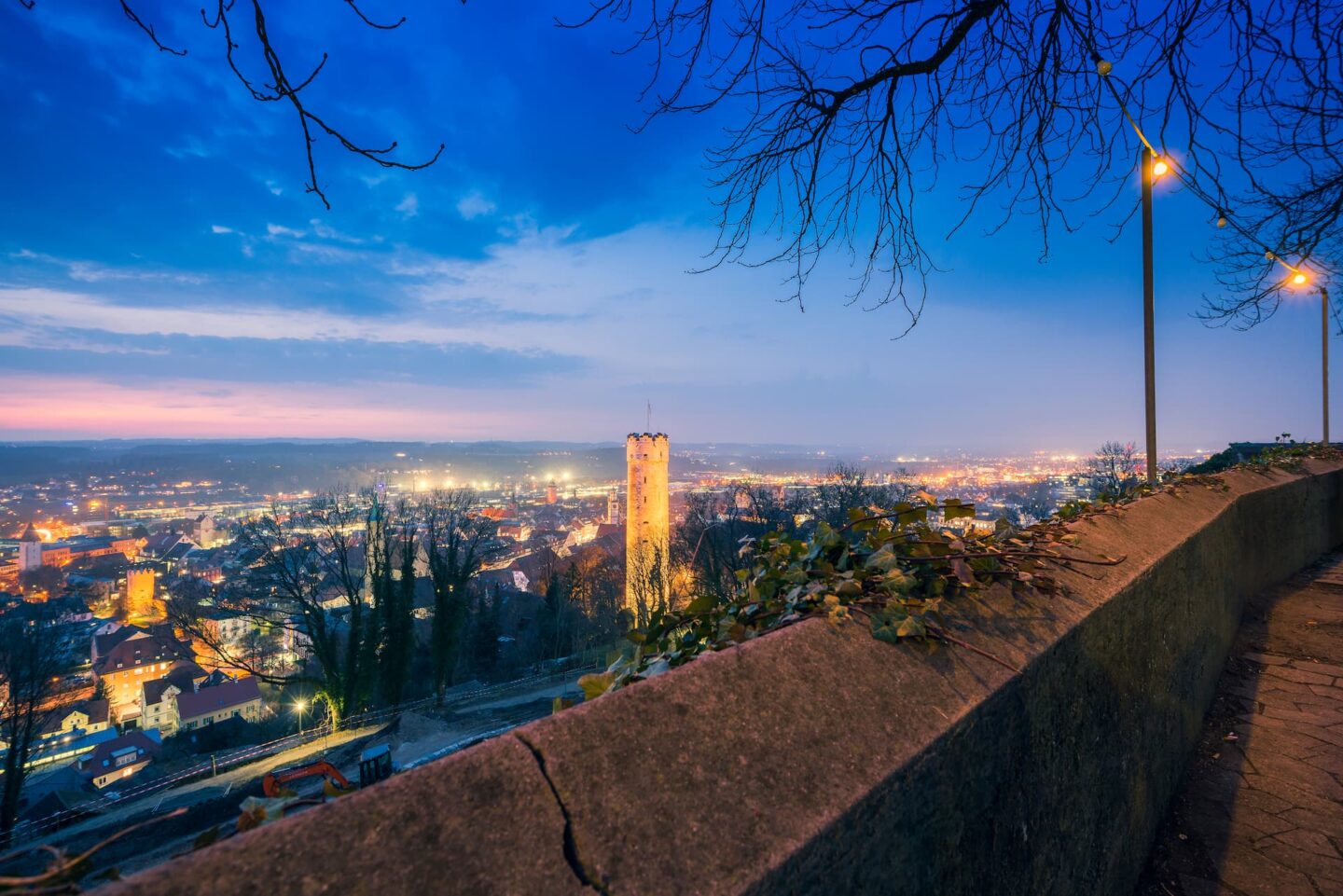 Panoramablick auf Ravensburg in der Abenddämmerung mit einem markanten Turm in der Mitte. Der Himmel ist in Blau- und Rosatönen gehalten und die Lichter der Stadt leuchten. Im Vordergrund sind eine Steinmauer und blattlose Äste zu sehen, die in die Szene hineinragen.