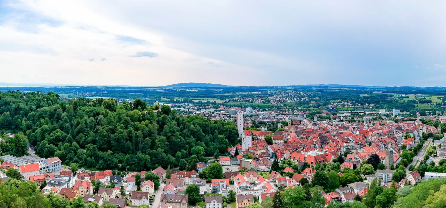 Ein Panoramablick auf Ravensburg zeigt eine Stadt mit einer Mischung aus roten und weißen Dächern, eingebettet zwischen grünen Bäumen und sanften Hügeln. Die Stadt erstreckt sich weitläufig, mit einem bewaldeten Hügel auf der linken Seite und einem eher wohnlichen Gebiet, das sich unter einem teilweise bewölkten Himmel in die Ferne erstreckt.