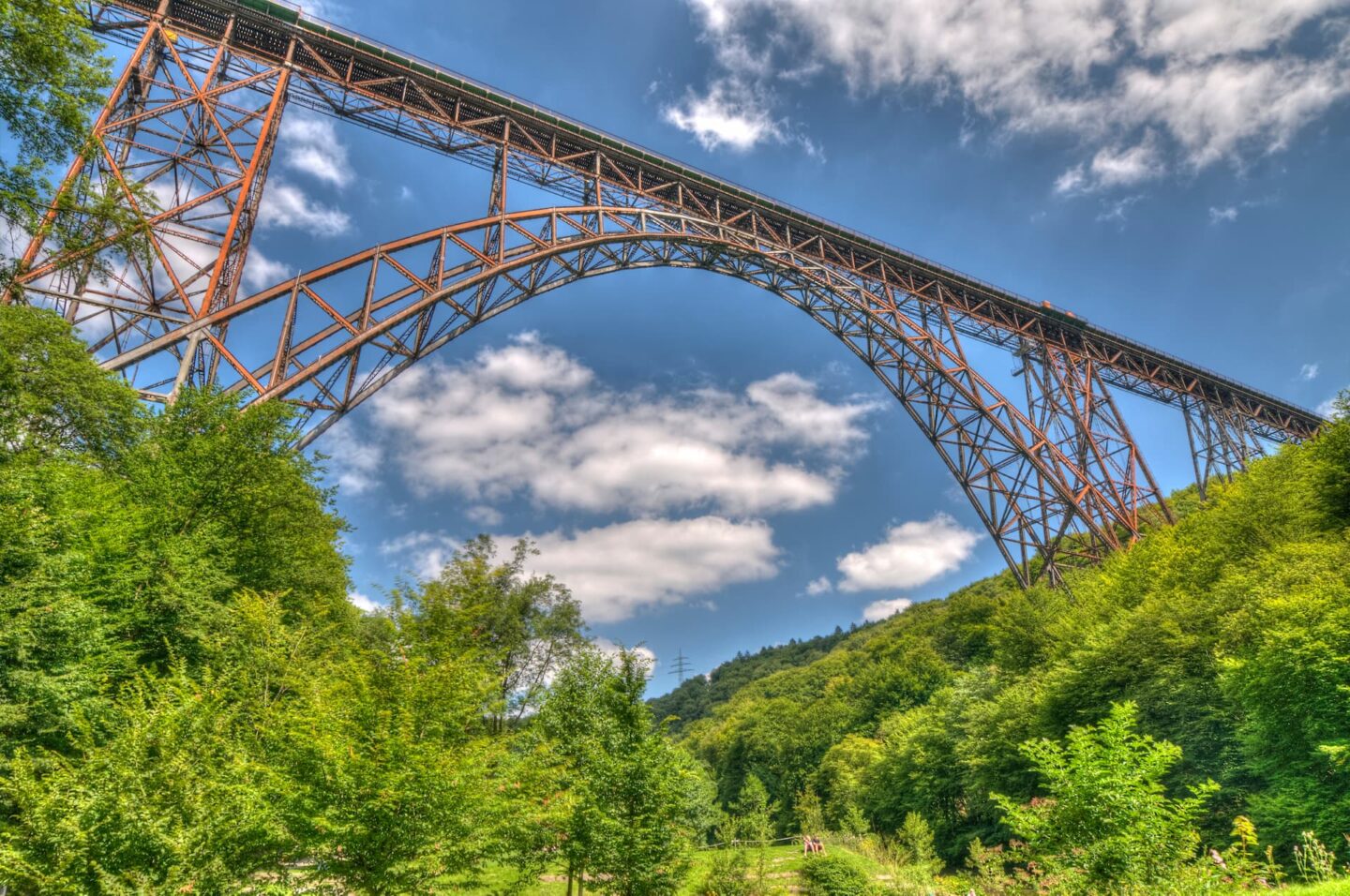 Ein Bild einer hohen, rostfarbenen Stahlbogenbrücke in Solingen, die sich über ein üppig grünes Tal spannt. Der Himmel darüber ist strahlend blau mit vereinzelten weißen Wolken. Bäume dominieren die Landschaft unterhalb der Brücke.