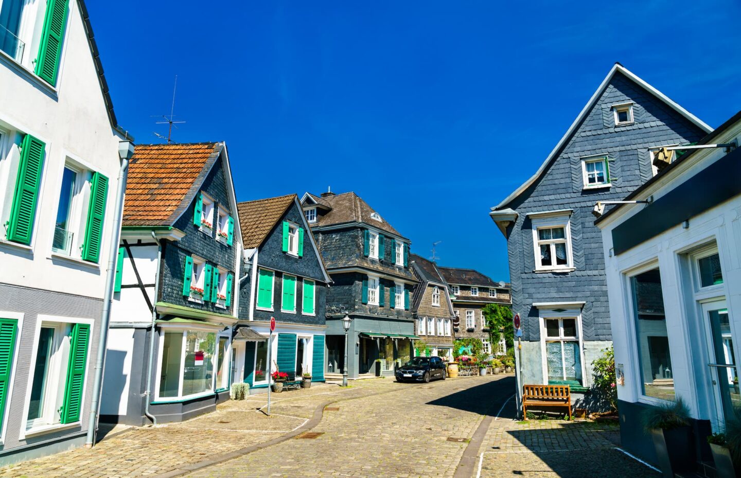 Eine malerische Kopfsteinpflasterstraße im europäischen Dorf Solingen, gesäumt von charmanten, farbenfrohen Häusern. Die Häuser haben Fensterläden und steile Dächer unter strahlend blauem Himmel. Auf dem Bürgersteig stehen eine Bank und Topfpflanzen, während in der Nähe ein Auto in dieser berühmten „Klingenstadt“ geparkt ist, die für ihre deutschen Messer berühmt ist.