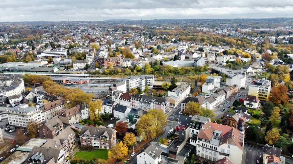 Luftaufnahme von Solingen, die einen Vorort mit zahlreichen Häusern, Gebäuden und Bäumen zeigt, die mit Herbstlaub geschmückt sind. Straßen und Wege kreuzen sich, während eine Eisenbahnlinie mit Zügen durch die Szene fährt. Der bewölkte Himmel bildet den Hintergrund für die ausgedehnte Landschaft.