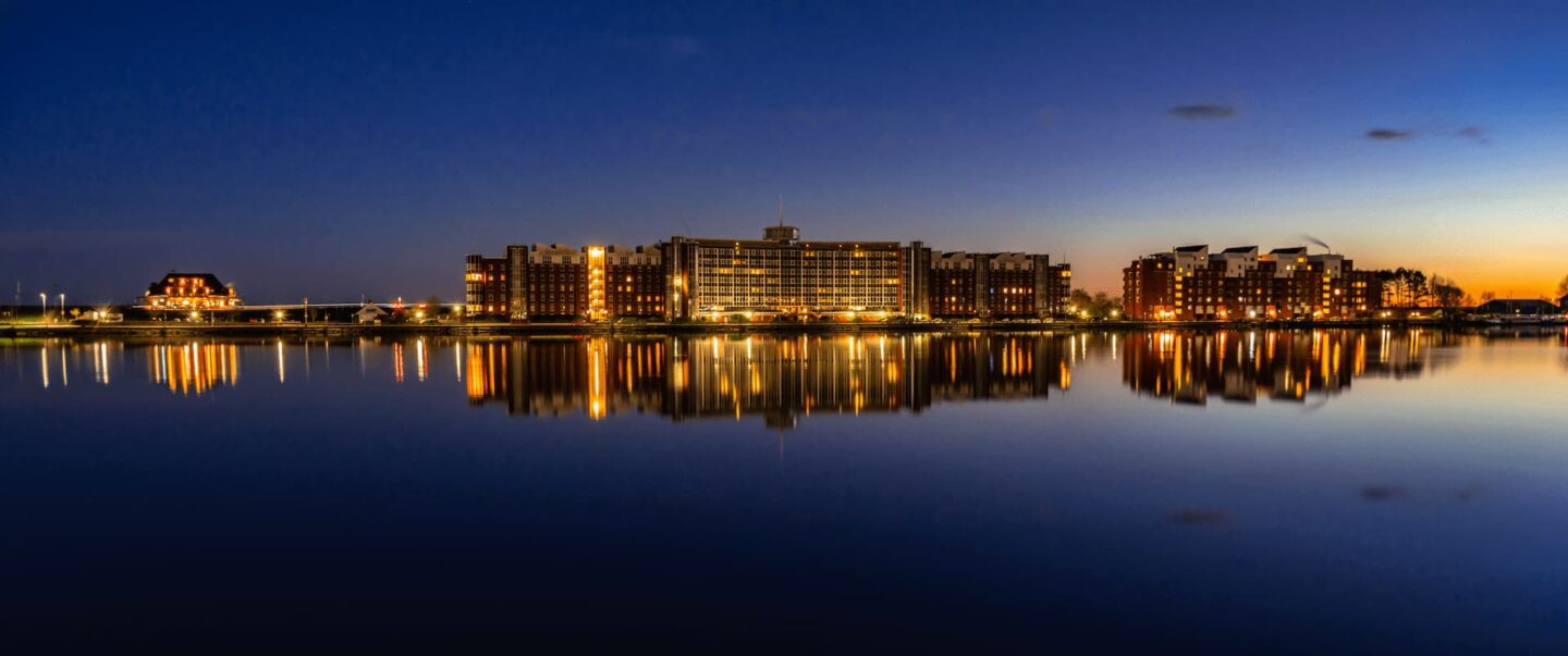 Eine Hafenlandschaft in Wilhelmshaven bei Abenddämmerung mit beleuchteten Gebäuden, die sich im ruhigen Wasser spiegeln. Der Himmel wechselt von einem tiefen Blau zum letzten Tageslicht. Straßenlaternen und Fenster erzeugen einen warmen Schein entlang der Uferlinie.