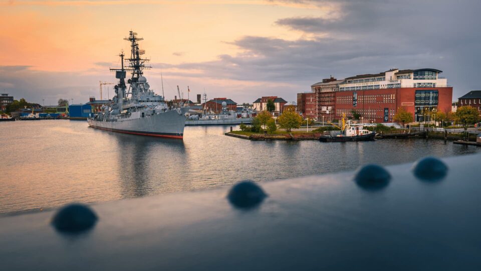 Ein großes Schlachtschiff ankert bei Sonnenuntergang in der Nähe der Stadtküste von Wilhelmshaven. Das ruhige Wasser spiegelt das Schiff und die Gebäude wider, im Hintergrund ist eine Mischung aus moderner und historischer Architektur zu sehen. Ein kleiner Schlepper ist in der Nähe. Der Himmel ist eine Mischung aus Orange- und Grautönen.