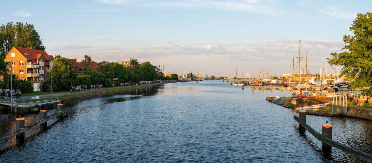 Panoramablick auf eine Szene am Flussufer in Wilhelmshaven mit angedockten Segelbooten auf der rechten Seite, einer von Bäumen gesäumten Promenade mit Menschen auf der linken Seite und einer Mischung aus roten Dächern und modernen Gebäuden im Hintergrund unter einem teilweise bewölkten Himmel.