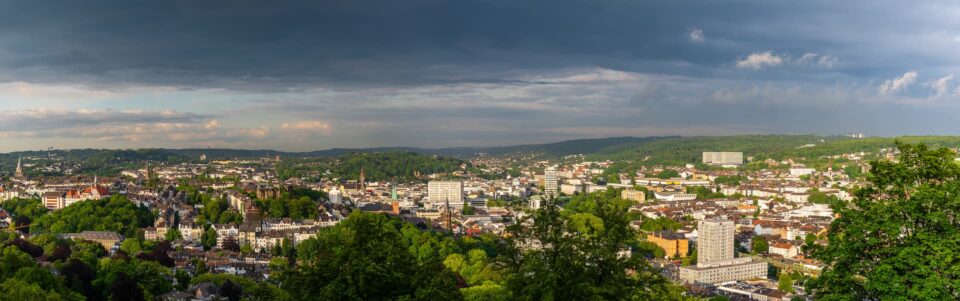 Panoramablick auf die Wuppertaler Stadtlandschaft mit einer Mischung aus modernen und traditionellen Gebäuden, umgeben von üppigen grünen Wäldern und Bäumen. Der Himmel darüber ist teilweise bewölkt, das Sonnenlicht bricht durch und wirft einen warmen Schein über die Stadt. In der Ferne erstrecken sich Hügel.