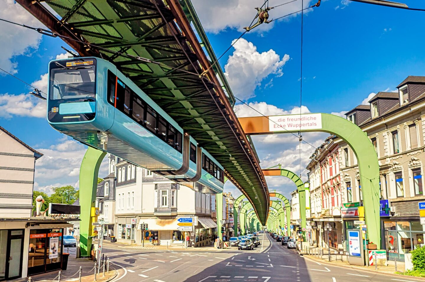 Eine leuchtend blaue Schwebebahn gleitet auf grünen, gewölbten Schienen über einer belebten Wuppertaler Straße. Auf beiden Seiten der Straße stehen Gebäude, darunter Autos und Fußgänger. Der Himmel ist blau mit vereinzelten weißen Wolken. Auf dem Schild an der Schwebebahn steht „die freundliche Elbe Wuppertals“.