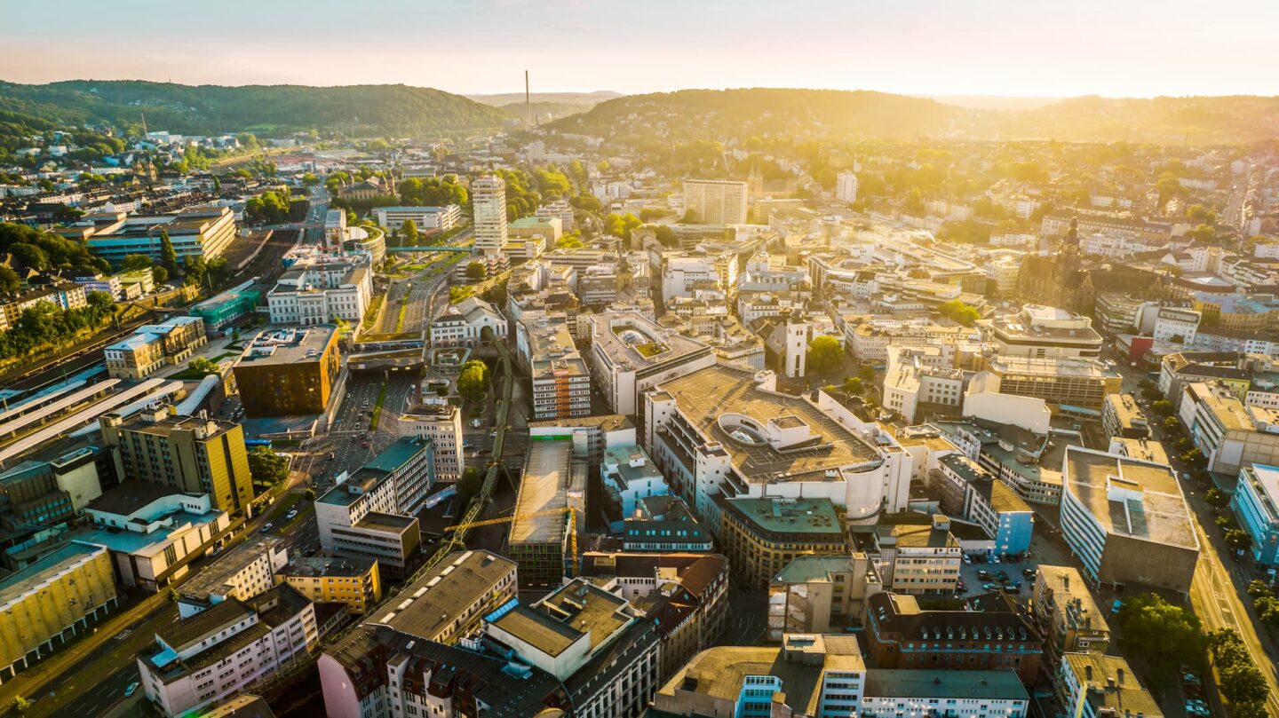 Luftaufnahme einer weitläufigen Stadtlandschaft bei Sonnenuntergang in Wuppertal. Die Szene zeigt eine Mischung aus modernen und traditionellen Gebäuden, Straßen und grünen Hügeln im Hintergrund. Die Sonne wirft ein warmes, goldenes Licht auf die gesamte Gegend und erzeugt lange Schatten und eine ruhige Atmosphäre.