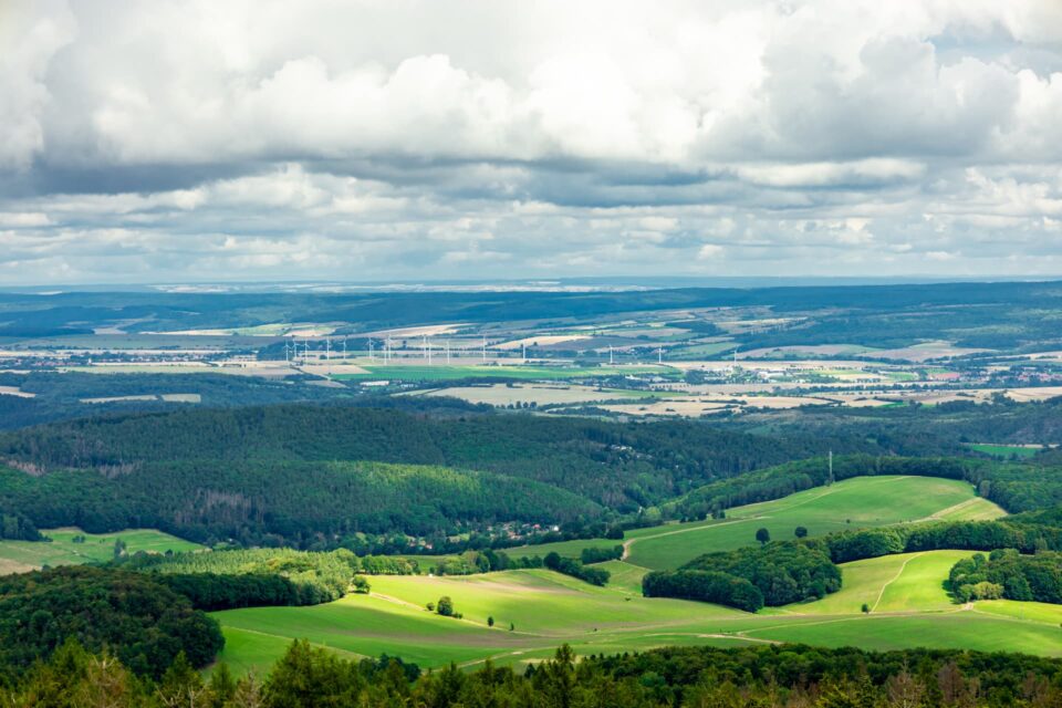 Eine malerische Landschaft in der Nähe von Wutha zeichnet sich durch sanfte grüne Hügel mit vereinzelten Bäumen und weitläufigen Feldern aus. Im Tal sind unter einem teilweise bewölkten Himmel Windturbinen zu sehen, die gesprenkeltes Licht auf das abwechslungsreiche Gelände werfen.