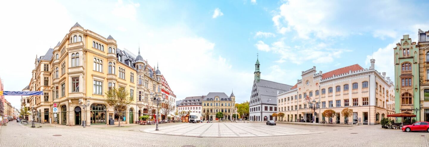 Panoramablick auf einen historischen europäischen Stadtplatz in Zwickau mit kunstvollen, mehrstöckigen Gebäuden in traditioneller Architektur. Der Platz ist offen und geräumig, mit Kopfsteinpflaster, ein paar Bäumen und einem zentralen Glockenturm. Der Himmel ist teilweise bewölkt.