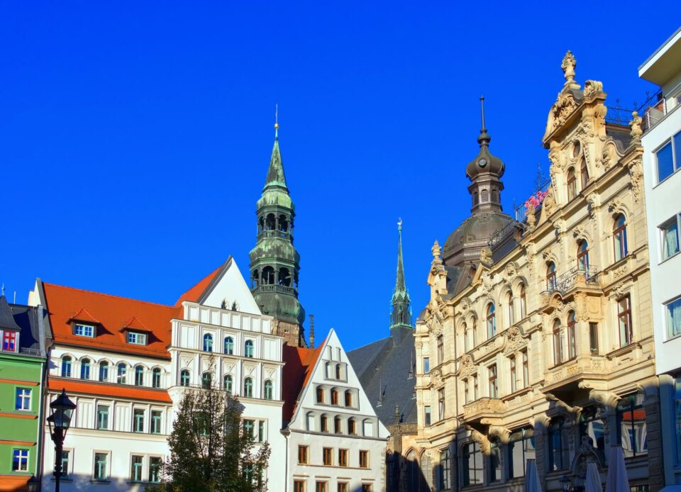 Eine malerische Stadtansicht von Zwickau mit historischen europäischen Gebäuden unter einem klaren blauen Himmel. Die Architektur weist unterschiedliche Stile auf, von kunstvollen klassischen Fassaden bis hin zu spitzen Dächern und Türmen, darunter ein markanter Uhrturm im mittleren Hintergrund.