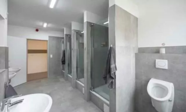 Badezimmer in einer Monteursunterkunft von Home4Time. Das Bad ist modern und mit grauen Fliesen ausgestattet. Es stehen mehrere Waschbecken und drei Duschen zur Verfügung. Rechts vorne im Bild sieht man ein Pissoir.