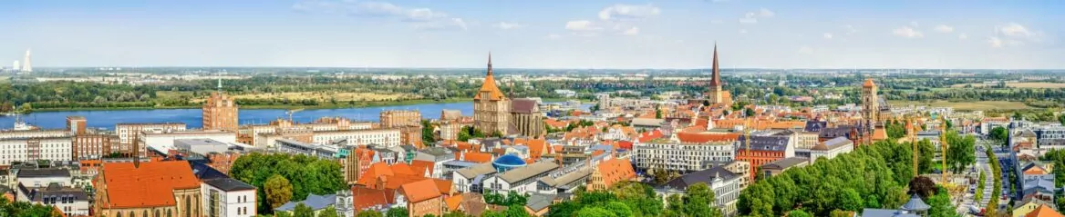 Ein Panoramablick auf eine europäische Stadt, durch die ein Fluss fließt. Das Stadtbild zeichnet sich durch zahlreiche Gebäude mit roten Dächern, Kirchtürme und Grünflächen aus. Im Hintergrund erinnern ausgedehnte Felder und ein klarer blauer Himmel mit einigen Wolken an den Charme Mecklenburg-Vorpommerns.