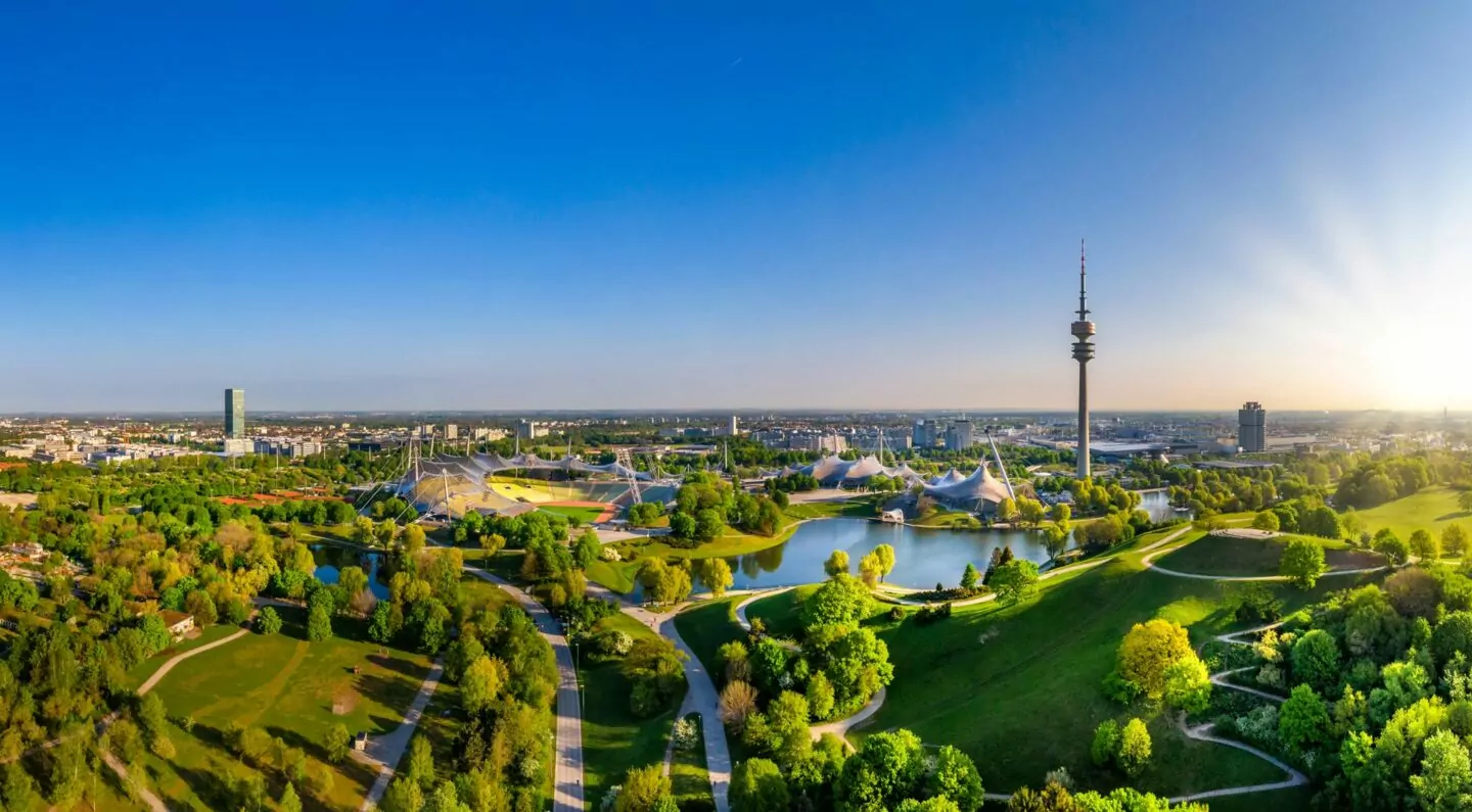 Ein Panoramablick auf die sonnendurchflutete Stadtlandschaft Münchens mit üppigen grünen Parks, einem großen See und einem markanten Turm. Die Skyline umfasst verschiedene Gebäude vor einem klaren blauen Himmel. Im Vordergrund sind Wege und Bäume zu sehen, die eine ruhige, städtische Szene bieten.