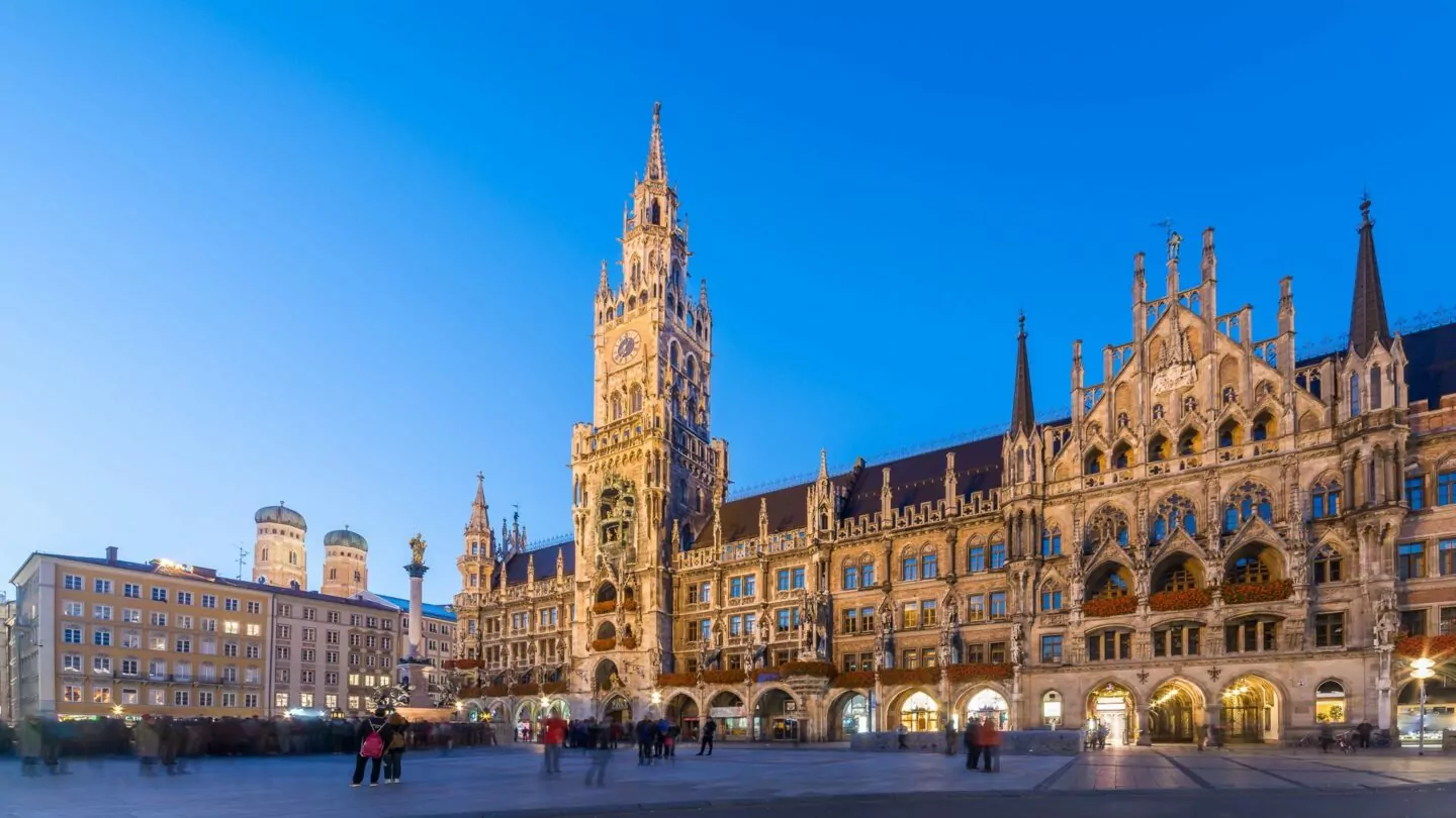 Eine lebhafte Abendszene am Marienplatz in München, Deutschland, mit dem beleuchteten neugotischen Neuen Rathaus. Der Turm und die kunstvolle Fassade des majestätischen Gebäudes heben sich vom klaren blauen Himmel ab. Die Menschen gehen spazieren und genießen die lebhafte Atmosphäre.