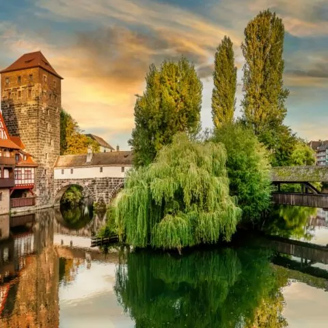 Ein malerischer Blick auf das historische Nürnberg bei Sonnenuntergang zeigt Fachwerkhäuser, einen Steinturm und üppiges Grün, das sich in einem ruhigen Fluss spiegelt. Eine Brücke überquert den Fluss und verbindet Teile der Stadt. Bäume und Gebäude vervollständigen die ruhige Szene.