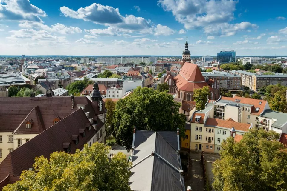 Ein Blick von oben auf die Stadt zeigt eine Mischung aus modernen und traditionellen Gebäuden unter einem klaren blauen Himmel mit vereinzelten Wolken. Inmitten dieses Cottbus-Panoramas sind zwischen den Gebäuden eine markante Kirche mit rotem Dach und verschiedene grüne Bäume zu sehen.