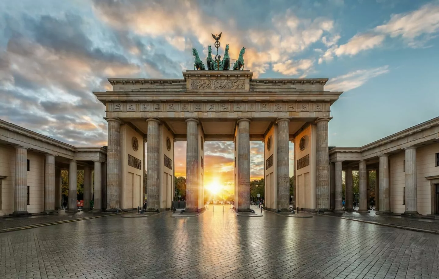 Das Brandenburger Tor in Berlin, Deutschland, beleuchtet von einem goldenen Sonnenuntergang. Dieses neoklassizistische Denkmal, ein Highlight des Städtetourismus, verfügt über hohe Säulen und eine Quadriga-Statue auf der Spitze, mit dramatischen Wolken im Hintergrund und einem ruhigen, nachdenklichen Innenhof im Vordergrund.