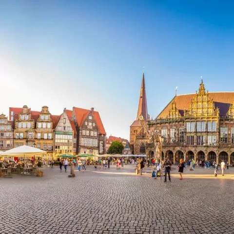 Panoramablick auf den historischen Stadtplatz von Bremen mit Kopfsteinpflaster, Straßencafés und flanierenden Menschen. Zu den umliegenden Gebäuden gehören eine gotische Kirche mit einem hohen Turm und verschiedene kunstvolle Gebäude mit roten Ziegeldächern und kunstvollen Fassaden vor einem klaren blauen Himmel.