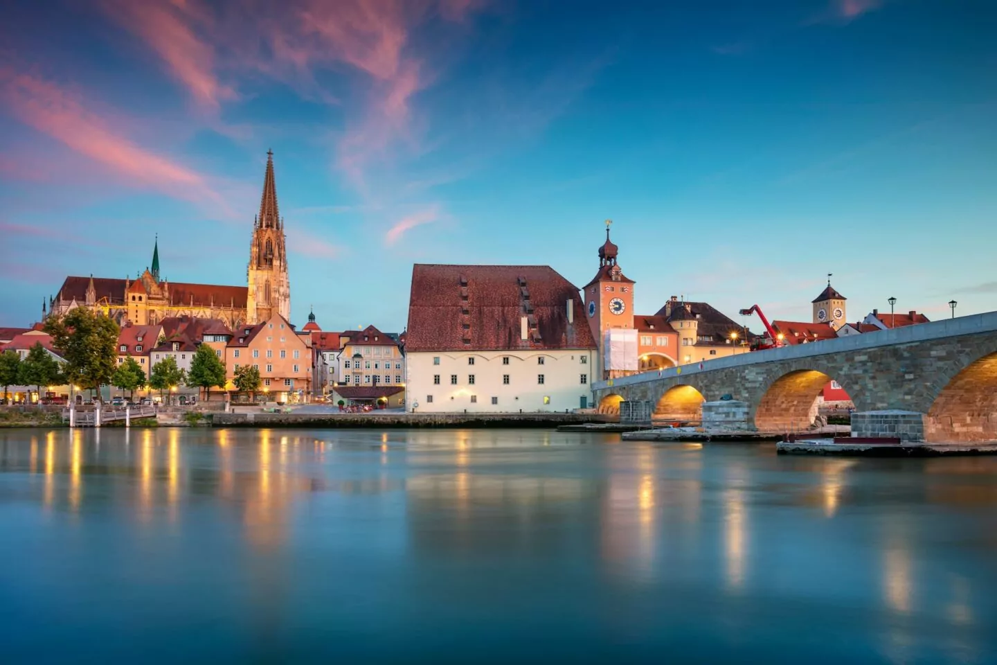 Ein ruhiger Blick auf Regensburg am Flussufer in der Abenddämmerung. Zu den markanten Merkmalen gehören eine große Kirche mit einem hohen Turm, historische Gebäude mit roten Ziegeldächern und eine gewölbte Steinbrücke. Der Himmel ist eine Mischung aus Blau- und Rosatönen, die sich im ruhigen Fluss spiegeln.